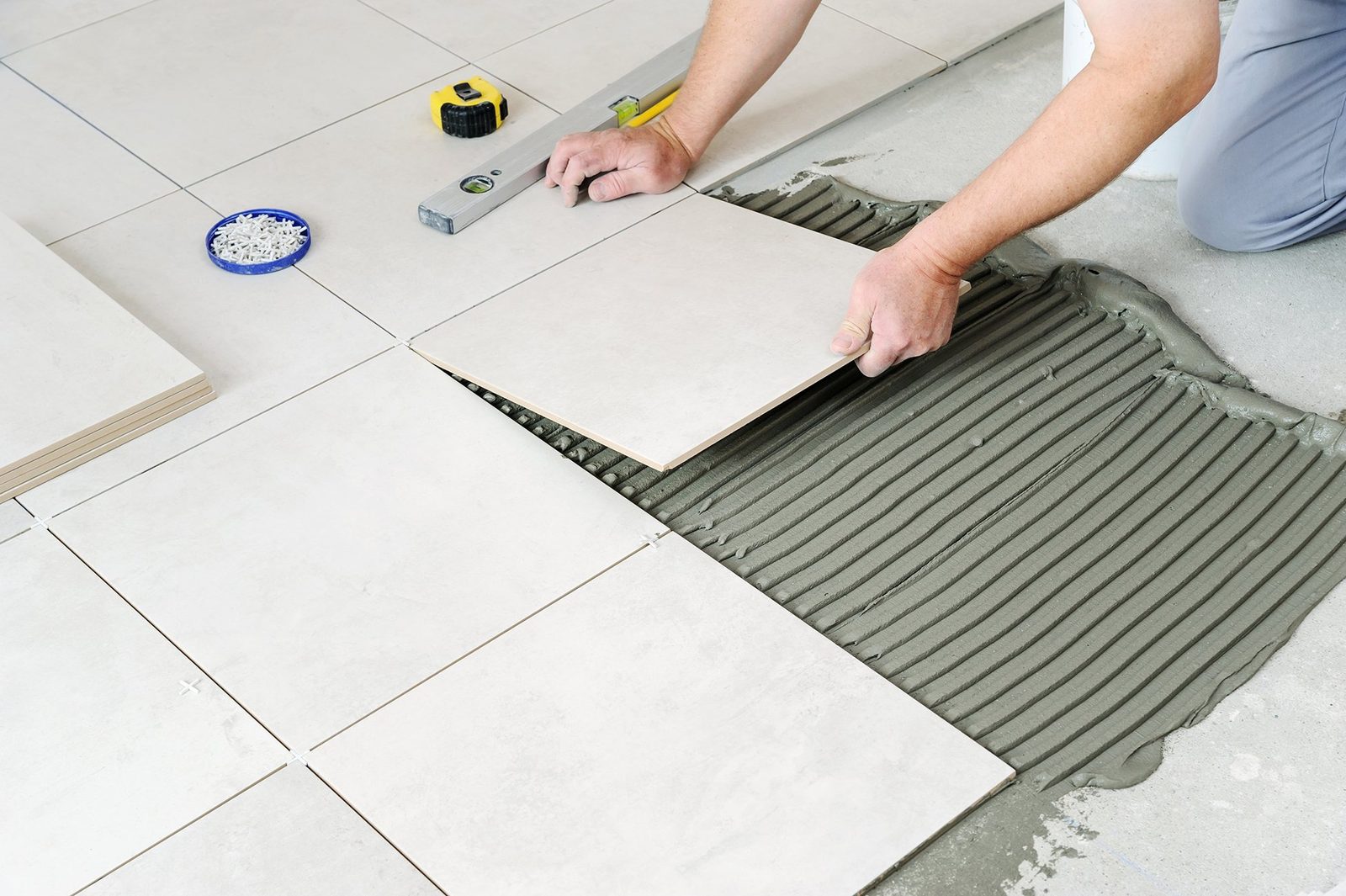 Installer setting a large floor tile into freshly troweled thin-set, with tile spacers and a level nearby